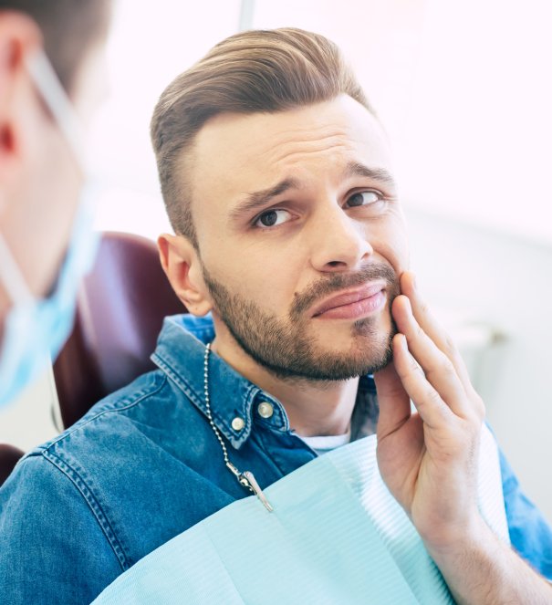 Symptoms of gums pain. A man with a worried face is holding his hand on his cheek because of irritating pain in front of a dentist who is going to give a patient a treatment. man smiling in dentists chair
